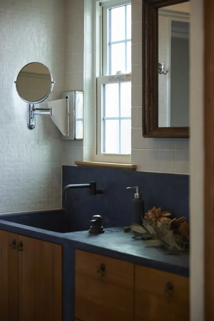 白いモザイクタイルの壁とモルタル調のカウンターが特徴的な、アンティクのモデルハウスの造作洗面台。無垢材のキャビネットやマットブラックの壁付け水栓、木枠のアンティーク調ミラーが備えられた、こだわりの洗面空間