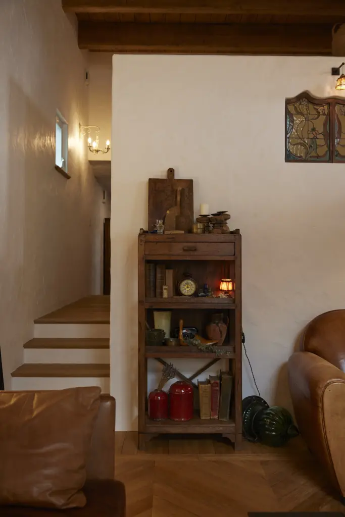 白い漆喰調の壁とヘリンボーン床の空間に配置された、アンティーク時計や古書などの小物がセンス良くディスプレイされている木製のオープンシェルフ。アンティクのモデルハウスの、こだわりが詰まったインテリア