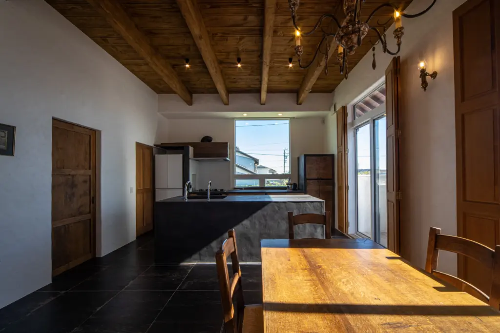 むき出しの梁がある板張り天井と黒いタイル床が特徴的な、アンティクのモデルハウスのダイニングキッチン。白い漆喰調の壁に、重厚感のあるアイランドキッチンやアンティーク家具が調和する、日当たりの良い空間