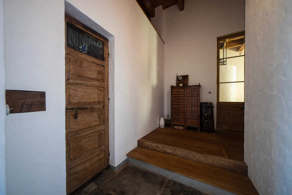 漆喰調の白い壁と無垢材の床が広がる、アンティクのモデルハウスの玄関。アイアン装飾が施されたアンティークの木製ドアや古いチェストが配置された、趣のあるエントランス空間