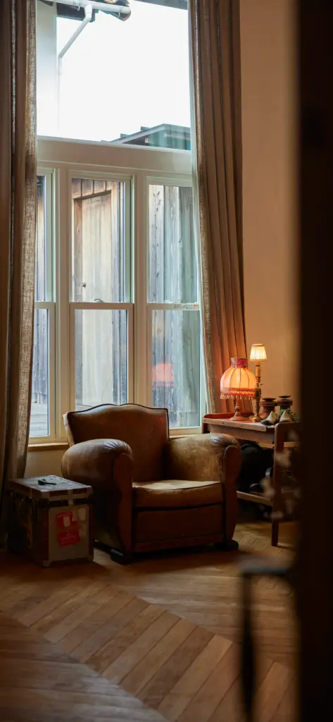 無垢材の床と大きな窓がある、アンティクのモデルハウスの窓際スペース。アンティークの1人掛けレザーソファや古いトランクケース、テーブルランプが配置されたリラックスできる空間