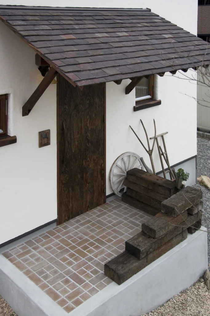 使い込まれた風合いの木製ドアと瓦屋根の庇が特徴的な、アンティクのA邸の玄関ポーチ。テラコッタ風タイルと枕木、古い車輪のディスプレイがアンティークな雰囲気を高めている
