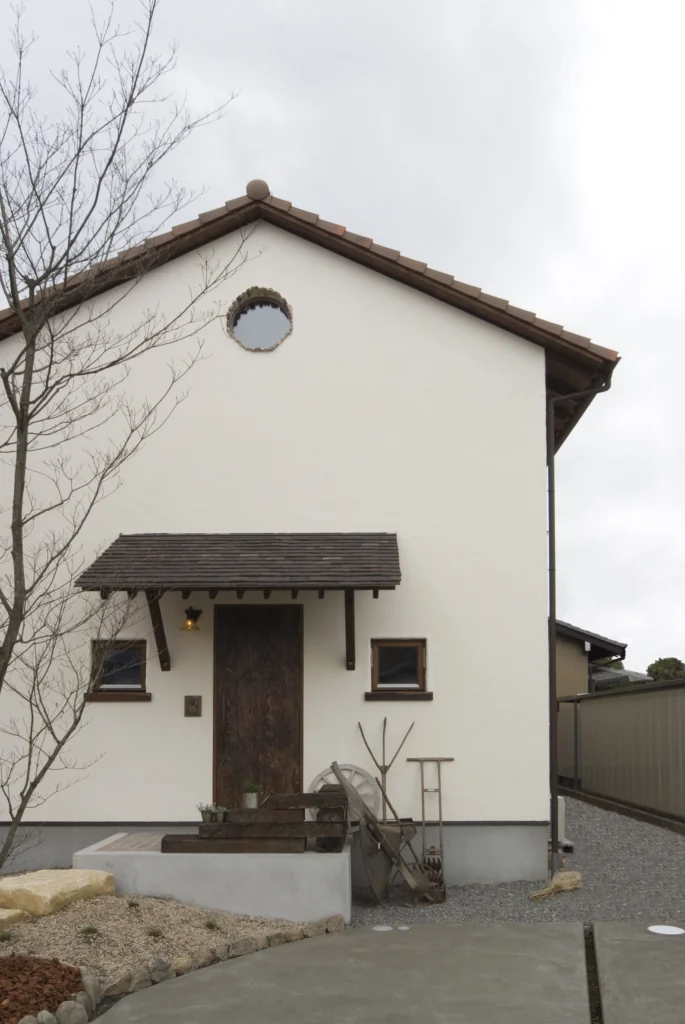 白い塗り壁に三角屋根、丸窓がアクセントになったアンティクのA邸の外観。枕木を使ったアプローチやアンティークな農具のディスプレイが、絵本から抜け出したような世界観を創り出している