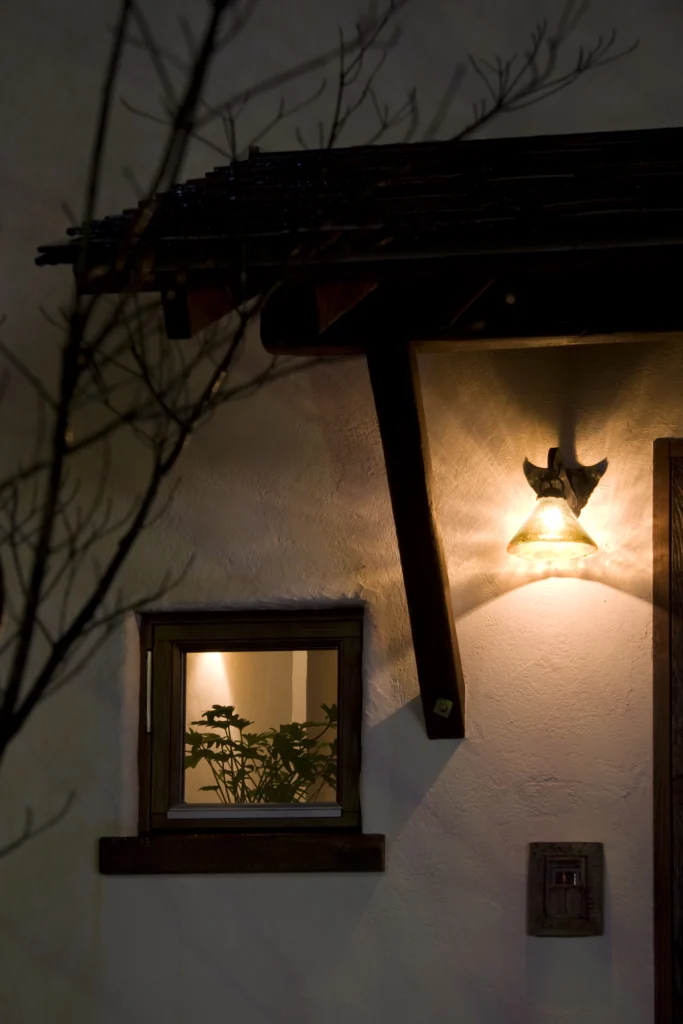 白い塗り壁に映えるアンティーク調のポーチライトが温かく出迎える、アンティクのA邸の夜の玄関ポーチ。小窓から漏れる光と植物のシルエットが幻想的な雰囲気を醸し出している