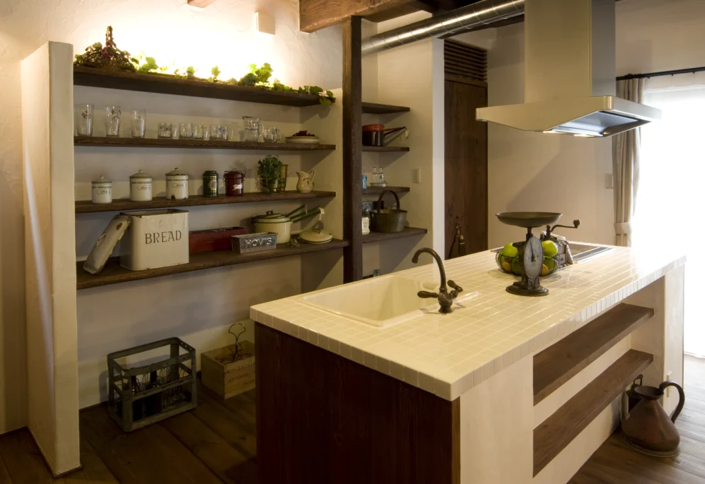 白いタイルの天板と木製のオープンシェルフが目を惹く、アンティクのA邸の造作アイランドキッチン。アンティーク調の水栓やディスプレイされた雑貨が、カフェのような空間を創り出している