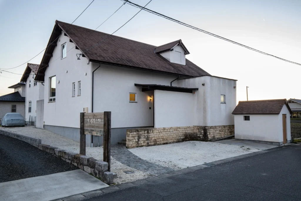 白い塗り壁と三角屋根が特徴的な、ANTiQのオフィス外観。手前にはアンティーク調の石積みの塀と木製看板が配置されている